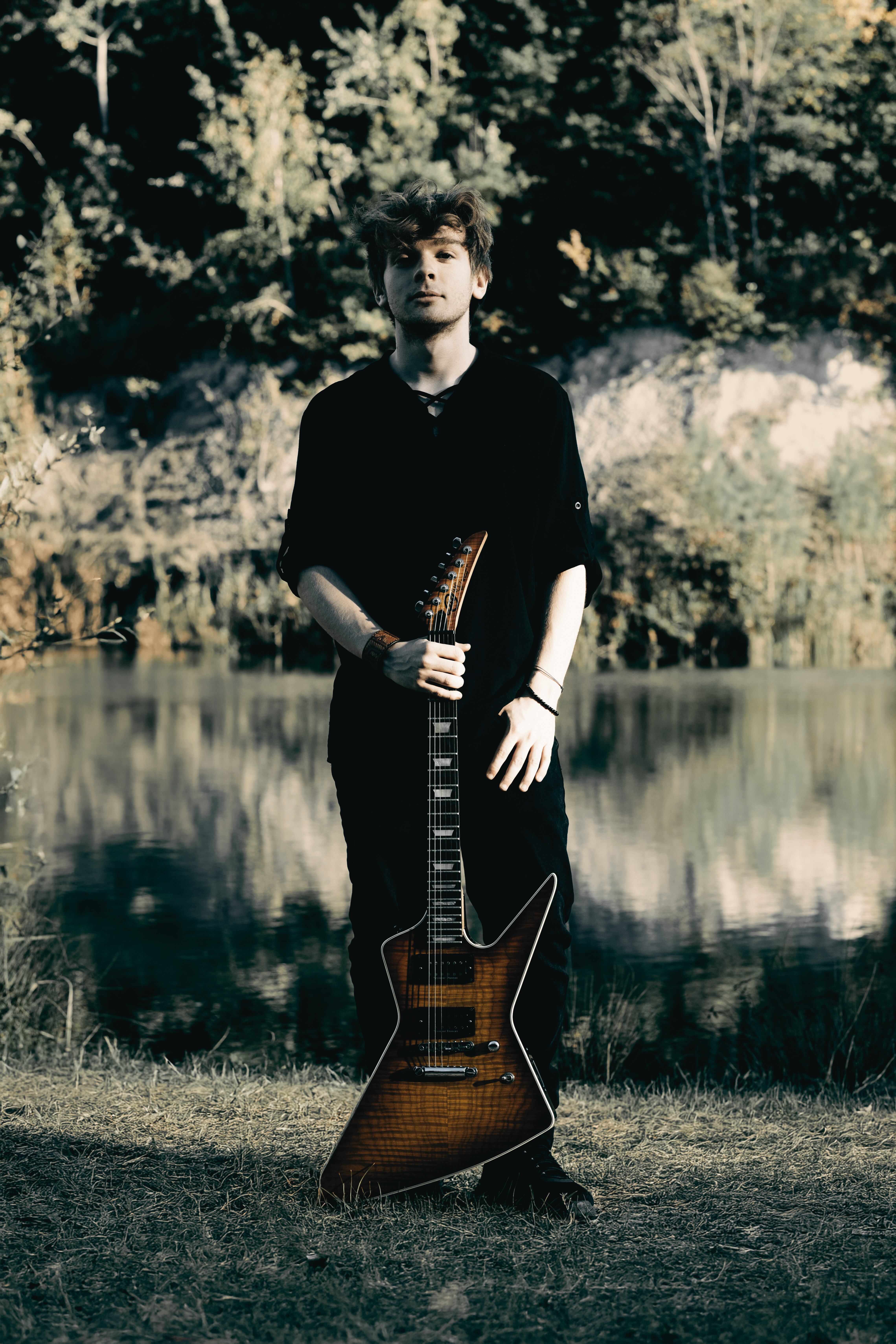 A young man with dark hair stands in a field, holding a guitar, with a serene lake and trees in the background.